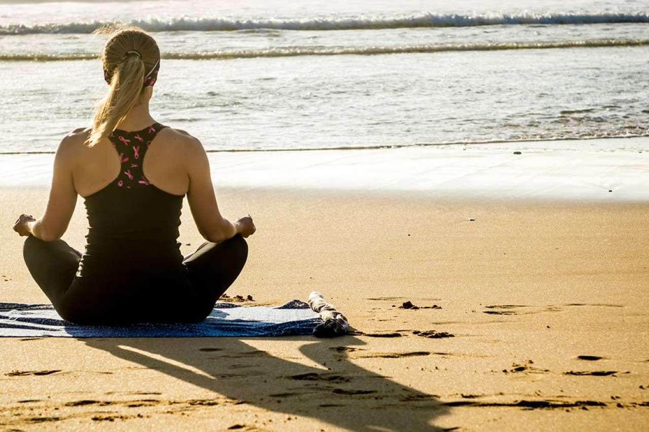 Woman meditating on a beach with her back towards the ocean 